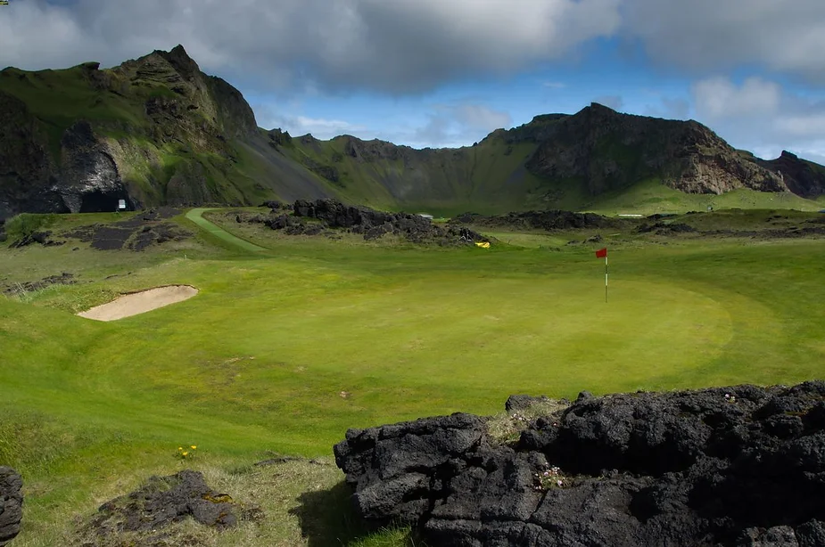 Scenic golf course in Iceland surrounded by rugged volcanic rock formations and lush green hills under a partly cloudy sky.