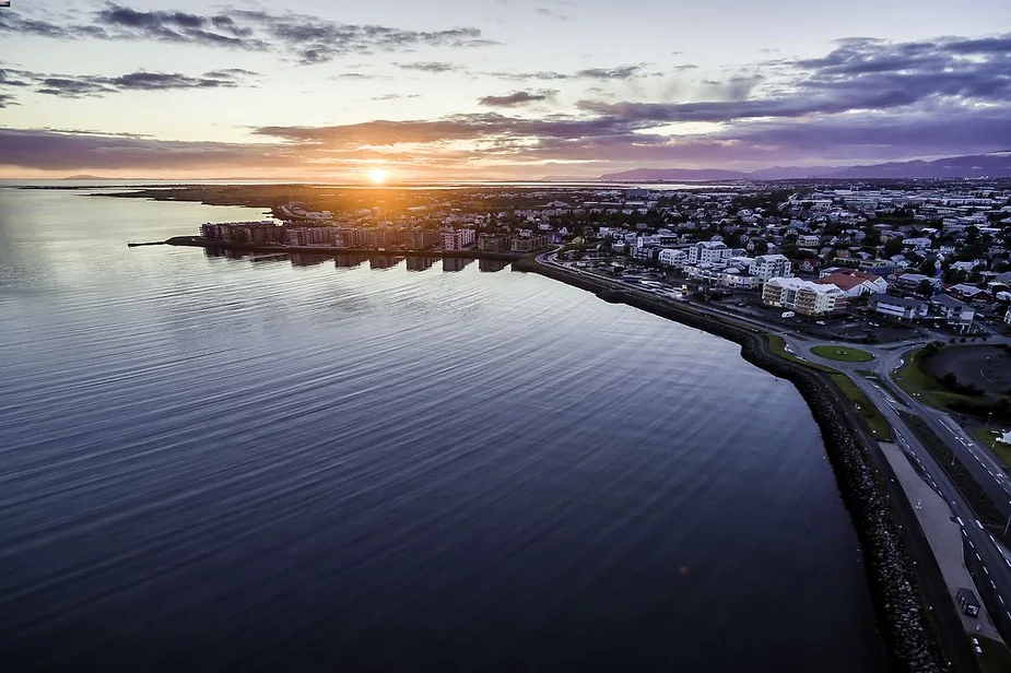 Aerial view of an Icelandic coastal town at sunset, with buildings and roads along the shoreline and a calm sea reflecting the setting sun.