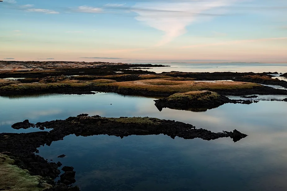 Serene coastal landscape in Iceland featuring calm waters, rugged rock formations, and a pastel-colored sky at dusk.