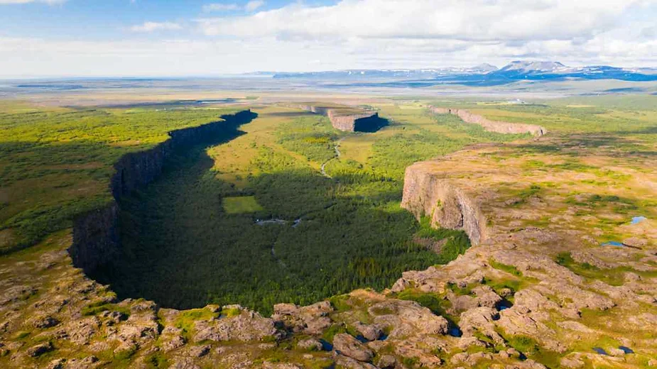 An aerial view of Asbyrgi Canyon in Iceland reveals a horseshoe-shaped, verdant valley surrounded by steep cliffs. The lush green vegetation contrasts with the rocky, barren landscape on the canyon's rim. The expansive view stretches towards distant mountains under a partly cloudy sky.