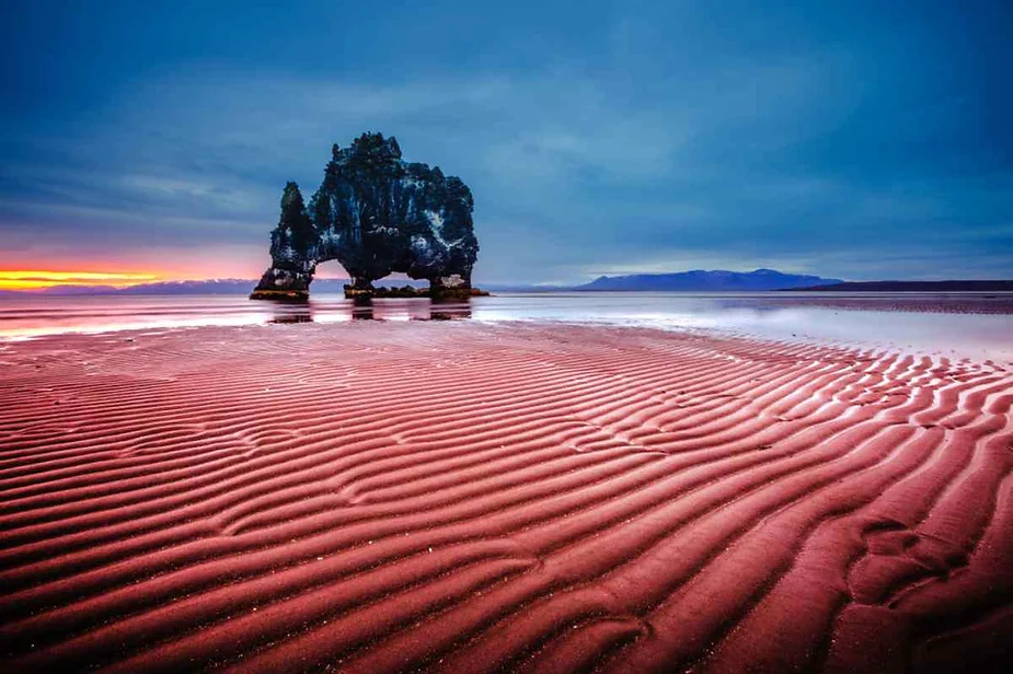 Hvítserkur, a distinctive rock formation, stands against a backdrop of a vibrant sunset on the shore of Iceland. The sand in the foreground features beautiful ripples, creating a textured pattern. The sky is a gradient of deep blues and purples, contrasting with the warm orange and yellow hues of the setting sun. The overall scene is tranquil and picturesque.