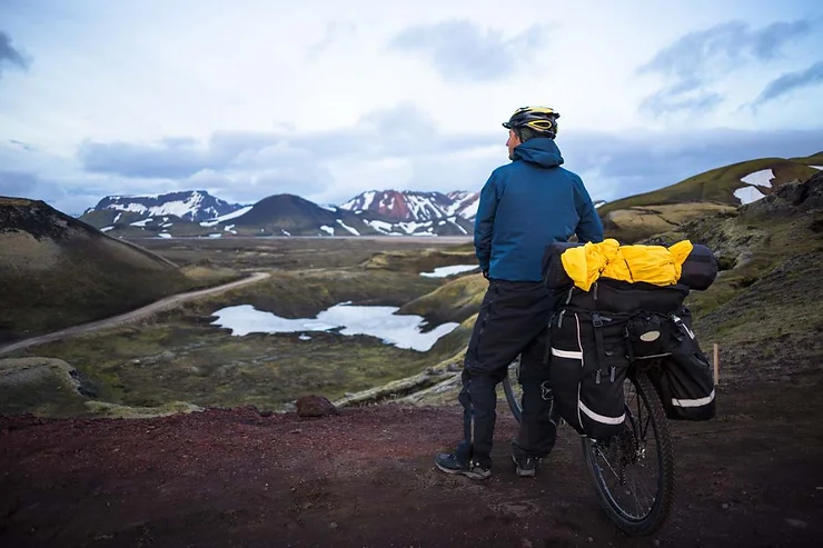 A cyclist standing beside their loaded bike, admiring the scenic landscape of Iceland with mountains and lakes in the background.