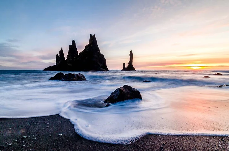 The stunning black sand beach in Iceland at sunset, with towering basalt sea stacks rising from the ocean in the background.