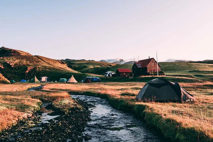 A serene campsite in Iceland featuring several tents set up near a small stream with a cabin and rolling hills in the background.