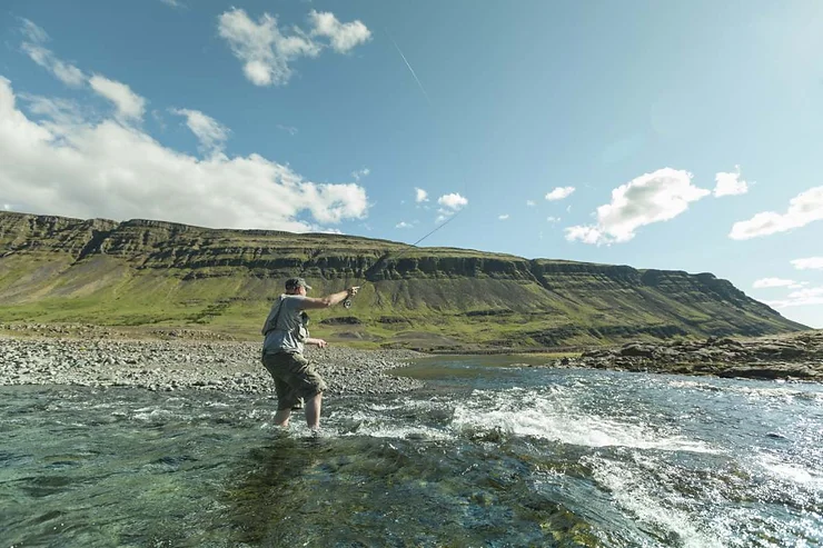 A person engaged in fly fishing in a clear, shallow river with a backdrop of majestic green hills under a bright blue sky with scattered clouds in Iceland.