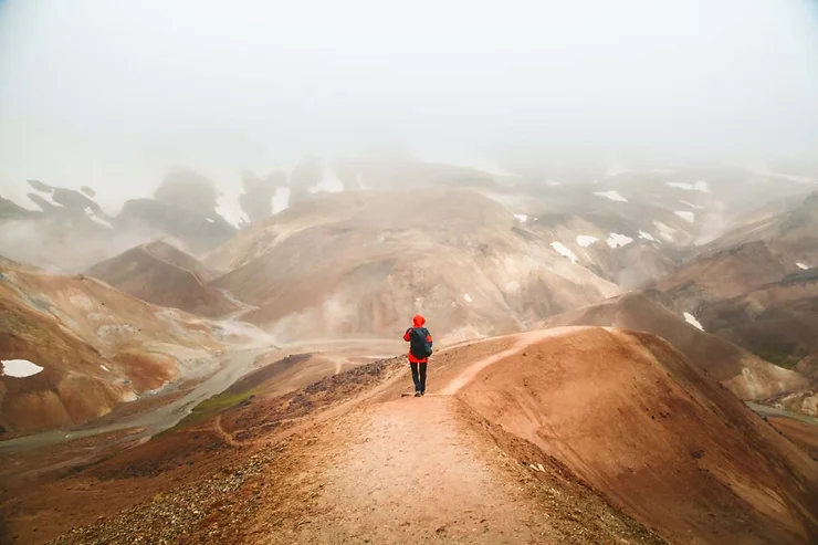 A lone hiker in red and blue gear walks along a trail through Iceland's rugged, foggy terrain with rolling hills and patches of snow.