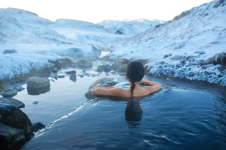 A person soaking in a hot spring surrounded by snow-covered rocks and mountains in Iceland. The warm water contrasts with the chilly winter landscape, creating a serene and relaxing atmosphere.