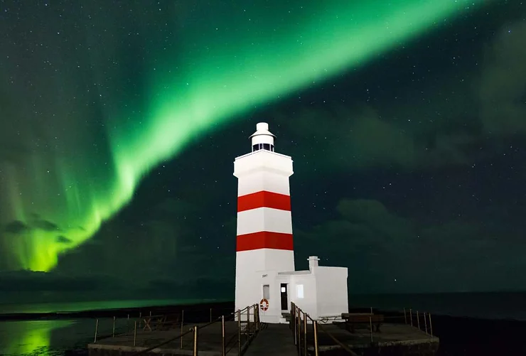 A white and red striped lighthouse illuminated by the stunning Northern Lights in Iceland.