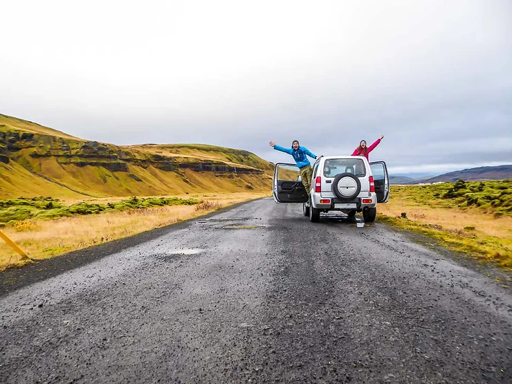 Two travelers with open car doors, joyfully stretching their arms out on a scenic road trip through Iceland's picturesque landscapes.