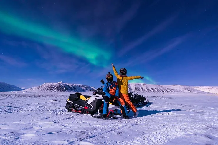 Two people on a snowmobile adventure in Iceland, posing with joy against a backdrop of snow-covered mountains and a mesmerizing display of the Northern Lights under a clear night sky.