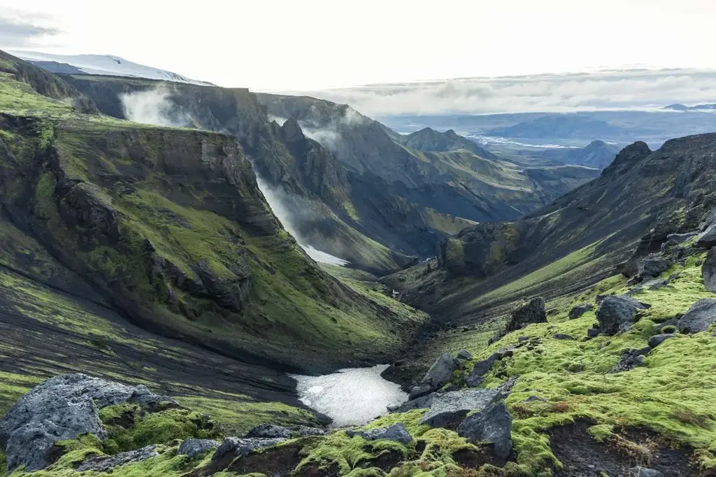 A dramatic view of the Fimmvörðuháls Trail in Iceland, featuring a rugged and steep valley draped in lush green moss and volcanic rocks. Mist rises from the depths of the valley, adding a mystical atmosphere to the scene. The trail winds through the landscape, showcasing the sharp contrasts between the dark, rocky slopes and the vibrant green vegetation. In the distance, the vast expanse of Iceland's highlands stretches out under a cloudy sky, inviting hikers to explore its challenging and breathtaking terrain.