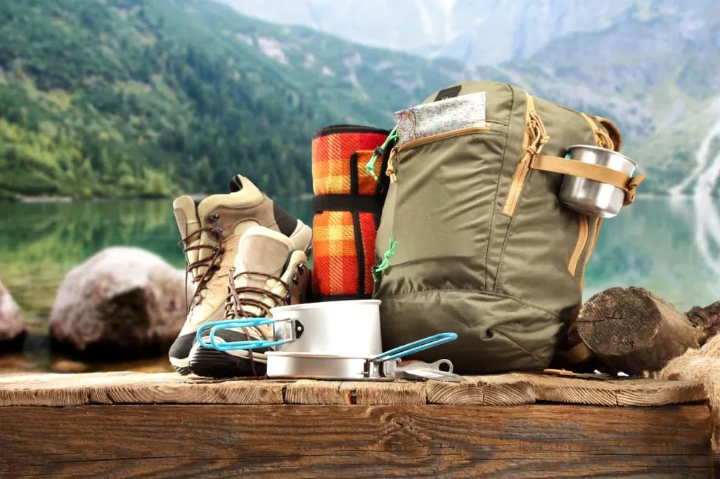 A set of essential hiking gear is neatly arranged on a wooden surface by a serene lake, surrounded by mountains in Iceland. The setup includes a sturdy pair of hiking boots, a green backpack with a metal cup attached, a rolled-up plaid blanket, and a portable camping stove with cooking utensils. This image captures the preparation needed for a comfortable and efficient hiking trip in Iceland's picturesque landscapes.