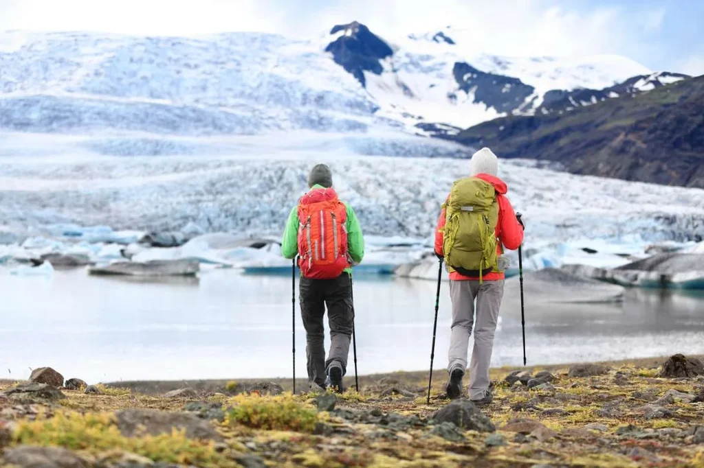 Two hikers, each equipped with trekking poles and backpacks (one orange and one green), walk towards a majestic glacier in Iceland. The expansive glacier, with its intricate ice formations and surrounding snow-capped mountains, provides a stunning backdrop. The terrain is a mix of rocky ground and sparse vegetation, highlighting the rugged beauty of Iceland's wilderness.