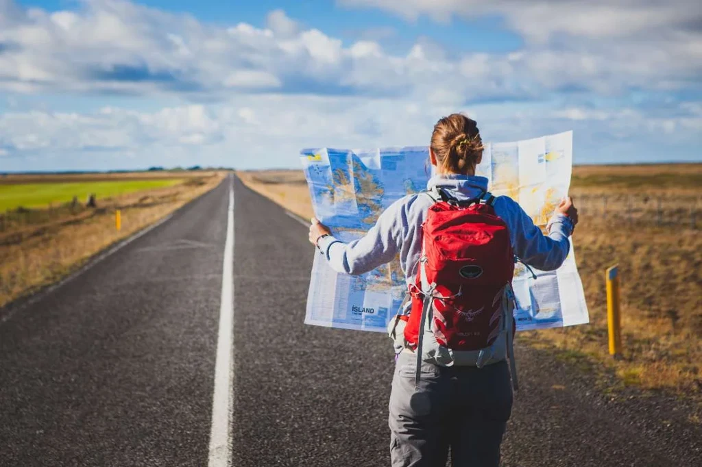 A hiker with a red backpack is standing on an open road in Iceland, holding a large unfolded map. The road stretches straight ahead into the horizon, flanked by fields and a vast open landscape under a partly cloudy sky. This image captures the spirit of adventure and exploration, highlighting the importance of navigation and preparation when hiking in Iceland.