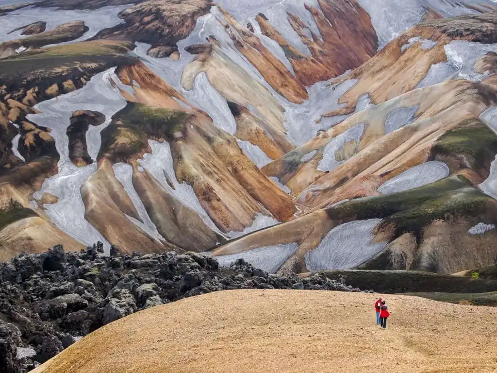 Two hikers, dressed in red jackets, stand on a sandy hill overlooking the strikingly colorful rhyolite mountains of Landmannalaugar, Iceland. The mountains are adorned with shades of orange, brown, and gray, creating a surreal, almost otherworldly landscape. The rugged terrain is interspersed with patches of green moss and dark volcanic rocks, adding to the dramatic scenery. This image captures the awe-inspiring beauty and unique geological features of Iceland's highlands, showcasing the adventurous spirit of hiking in this remote area.