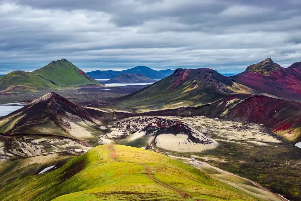 A stunning panoramic view of the Laugavegur Trail in Iceland, showcasing a vibrant landscape of multicolored mountains. The scene is dominated by hills and peaks in shades of green, red, and black, with patches of snow lingering in the crevices. The overcast sky casts a dramatic light over the rugged terrain, emphasizing the contrast between the lush moss-covered areas and the volcanic rocks. This image captures the breathtaking beauty and diverse geology of one of Iceland’s most famous hiking trails, inviting adventurers to explore its unique and scenic paths.