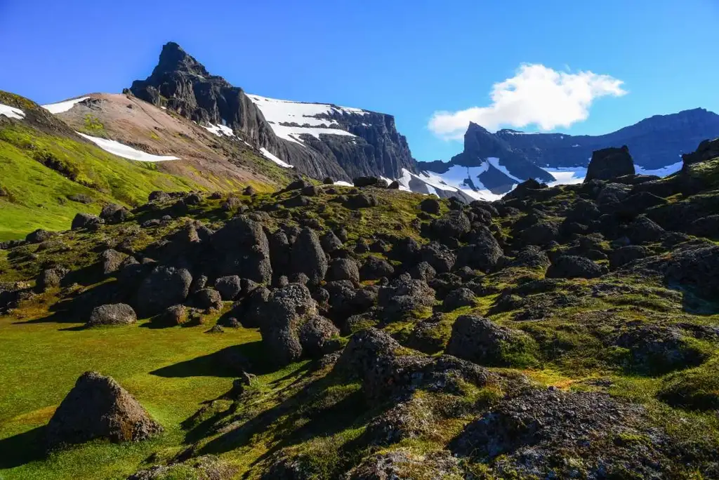 A stunning view of the Storurd Trail in Iceland, featuring rugged terrain dotted with large boulders and patches of vibrant green moss. In the distance, jagged mountain peaks rise against a brilliant blue sky, with traces of snow clinging to the slopes. The foreground is bathed in sunlight, creating a striking contrast between the dark rocks and the lush greenery. This dramatic landscape captures the raw, untouched beauty of Iceland’s highlands, offering a glimpse into the adventurous spirit of hiking this remote trail.