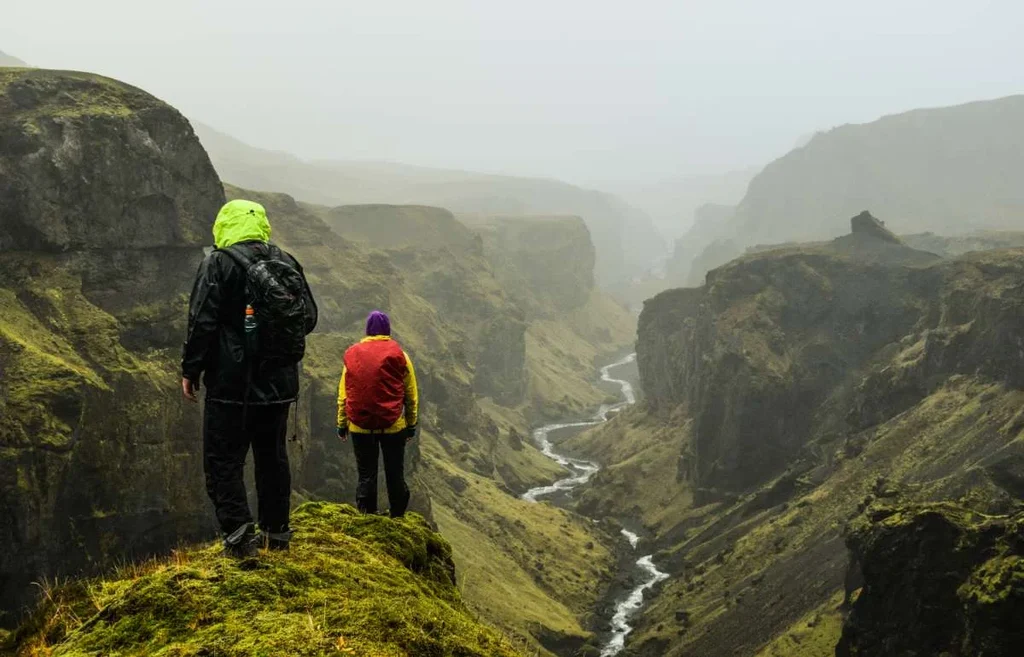 Two hikers stand on a misty cliffside overlooking a dramatic valley in Iceland. The foreground features the hikers in colorful, weatherproof gear, their backs facing the camera as they take in the sweeping view. The valley below is lush and green, with a river winding through the rugged terrain. The fog adds an ethereal quality to the scene, creating a sense of mystery and adventure in this remote and wild landscape.