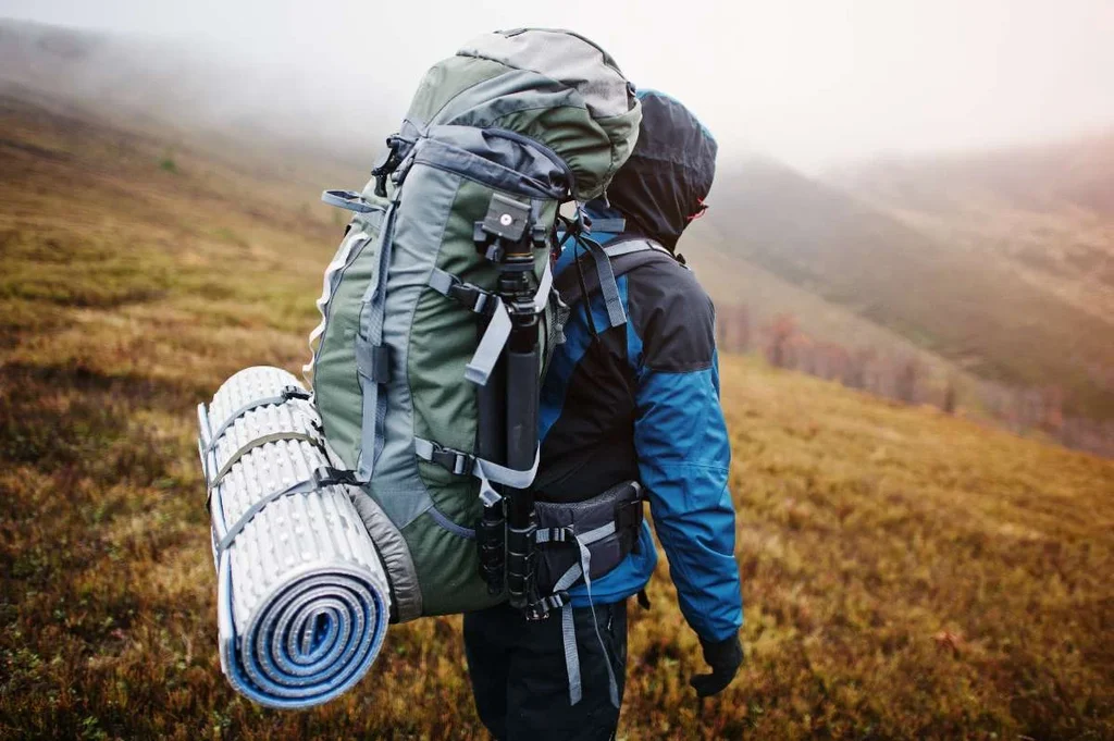 A hiker wearing a hooded jacket stands with their back to the camera, looking out over a misty, grassy landscape in Iceland. The hiker's large backpack is prominently visible, complete with a rolled-up sleeping mat attached to the bottom. The gear is essential for an extended hiking trip, indicating preparedness for the unpredictable Icelandic weather and terrain.