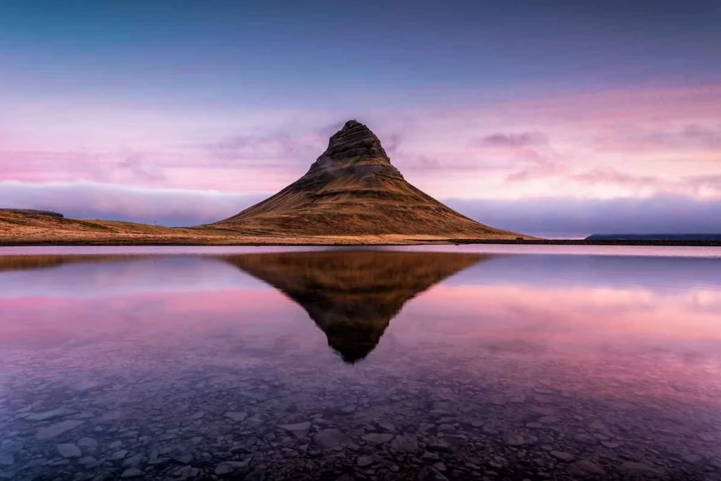 Kirkjufell mountain in Iceland, perfectly reflected in a calm body of water at sunset. The sky is painted with shades of pink, purple, and blue, casting a serene and almost surreal glow over the landscape. The mountain's distinct, symmetrical shape and the tranquil reflection create a mesmerizing scene.