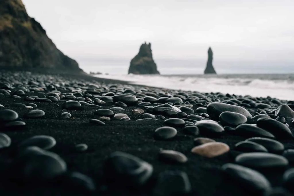 A close-up of smooth, black pebbles on Reynisfjara Beach in Iceland. The pebbles create a stark contrast against the black sand, with the dramatic basalt sea stacks rising in the background, shrouded in mist. The scene captures the raw and rugged beauty of this iconic Icelandic location.