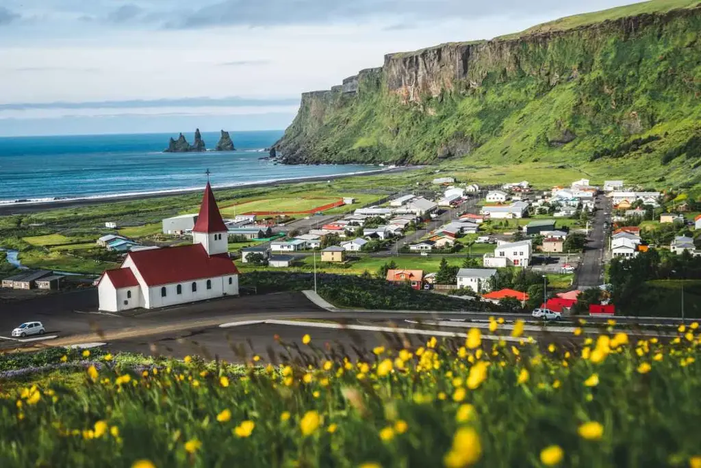 A picturesque view of Vik in Iceland, showcasing a charming village with colorful houses, a prominent white church with a red roof, and lush green cliffs in the background. The scene is framed by the vast blue ocean and the famous Reynisdrangar sea stacks. The foreground features a meadow dotted with yellow flowers, adding to the vibrant beauty of the landscape.