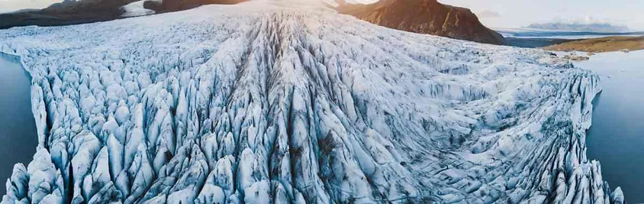 A breathtaking aerial view of a massive glacier in Iceland. The glacier's surface is a complex landscape of deep crevasses and jagged ice formations, stretching out toward the mountains in the background. The scene is bathed in soft sunlight, highlighting the pristine whiteness of the ice and the rugged beauty of the surrounding landscape. The glacier flows into a calm body of water on both sides, creating a striking contrast between the icy expanse and the serene waters.