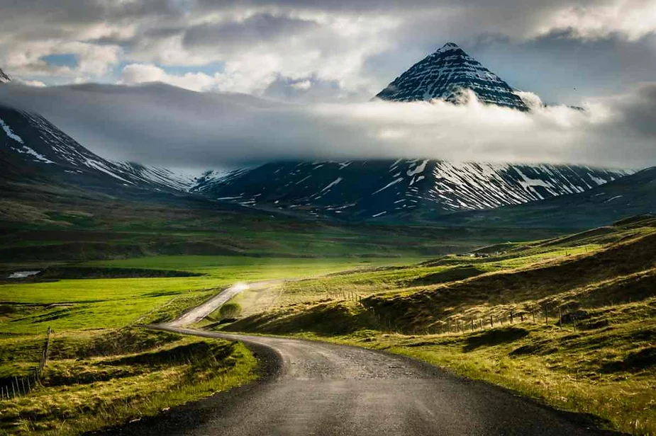 A winding gravel road leads through a lush green valley towards a majestic, pyramid-shaped mountain partially shrouded in clouds. The mountain's snow-capped peaks and the dramatic cloud cover create a striking contrast with the vibrant green pastures. The scene is serene and picturesque, capturing the rugged and tranquil beauty of Iceland's landscape.