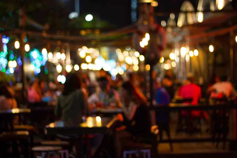 A bustling outdoor café at night, filled with people enjoying their evening. The image is characterized by a soft focus and warm bokeh effect, creating a dreamy atmosphere. String lights and lanterns illuminate the scene, casting a cozy glow on the patrons and the surroundings. The vibrant lights and the lively crowd contribute to the energetic and inviting ambiance of the café.