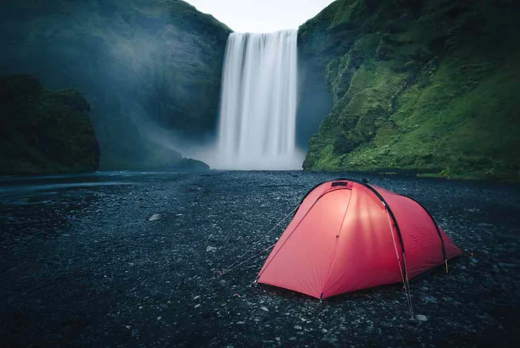 A red tent set up near the base of a tall waterfall surrounded by lush green cliffs in Skogar, Iceland.