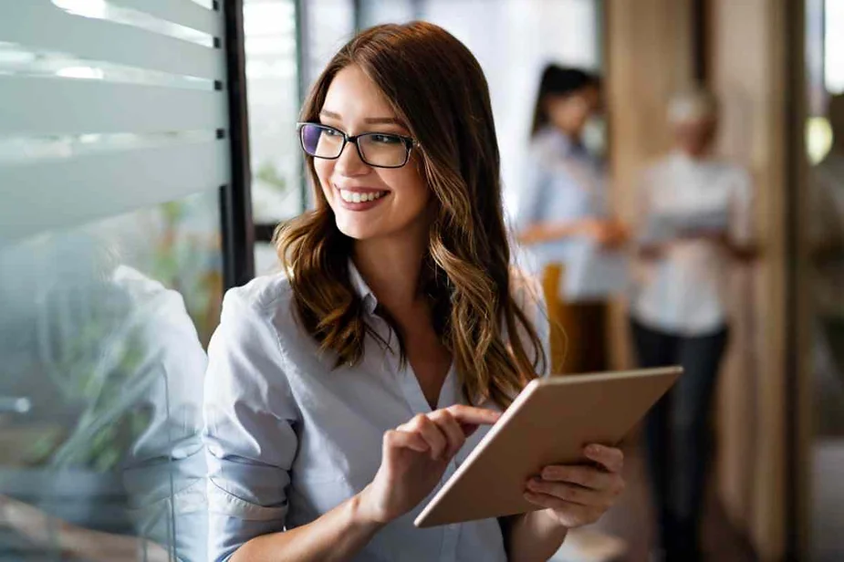 A smiling woman with glasses is holding and using a tablet. She is standing in an office environment with colleagues in the background.