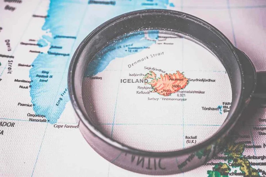 A map highlighting Iceland with a magnifying glass placed over it. The map shows the surrounding regions and bodies of water, with Iceland prominently focused in the center of the magnifying glass.