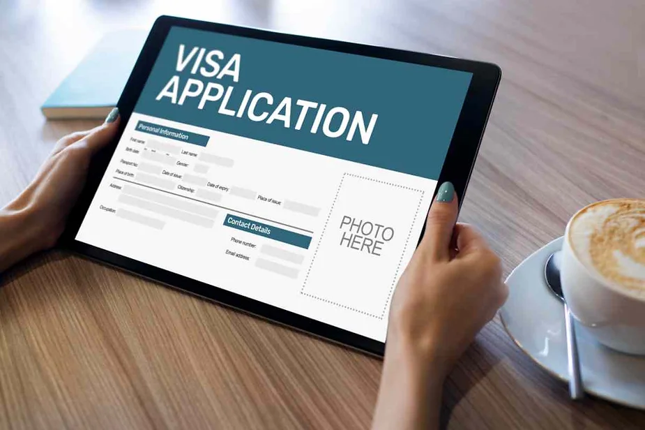 A person is holding a tablet displaying a visa application form. The screen shows fields for personal information and contact details, along with a placeholder for a photo. A cup of coffee is placed on the table beside the tablet.