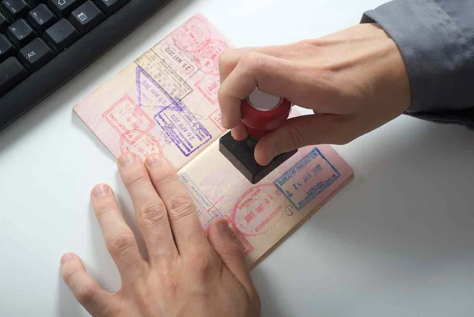 A person's hand stamping a passport with multiple visa and entry stamps visible. The scene includes a keyboard partially in view, suggesting a border control or customs office setting.