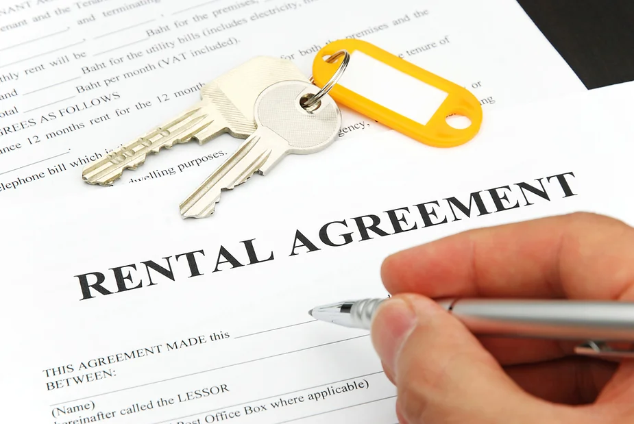 A hand holding a pen is signing a rental agreement document. The document is titled 