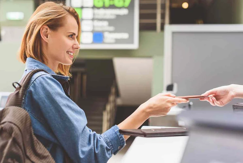 A young woman with a backpack is smiling as she hands over her passport to a security officer at an airport check-in counter. The background shows signs with gate information, indicating an airport setting.