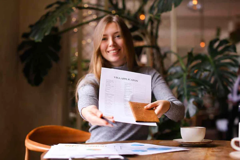 A smiling woman sitting at a table, holding out a visa application form and a passport, with a cup of coffee and documents on the table in front of her.