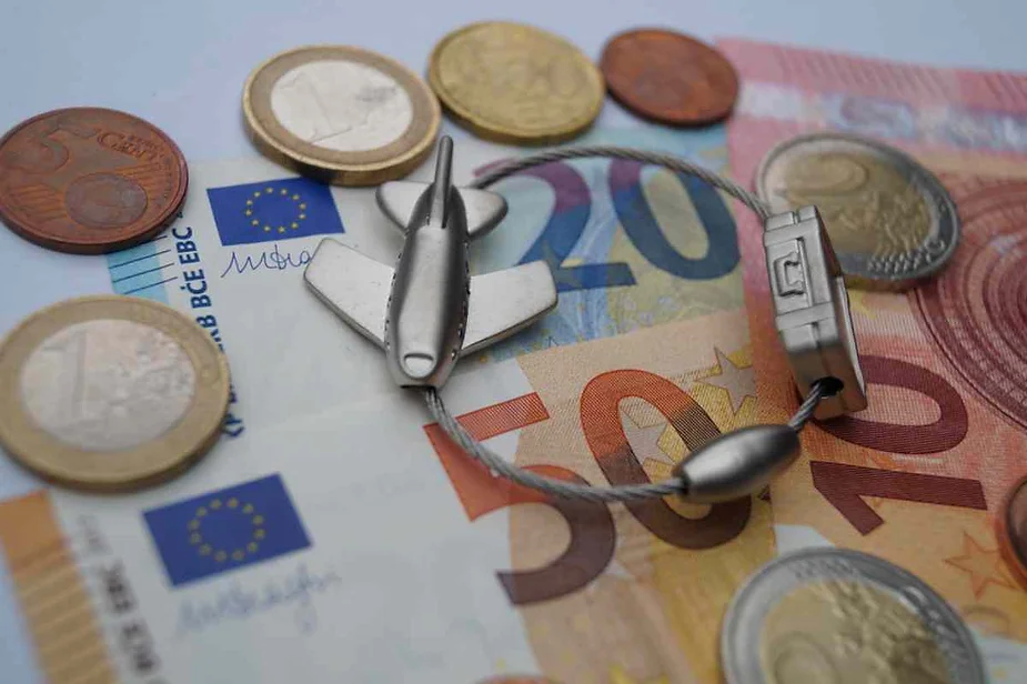 Various Euro banknotes and coins are spread out, featuring a keychain with a small airplane and luggage tag. The scene highlights the financial aspects of travel planning.