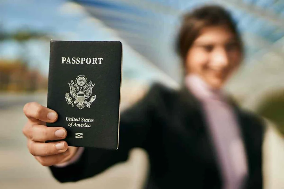 A person holds up a United States passport with a smile, showcasing it to the camera. The background is slightly blurred, highlighting the focus on the passport.