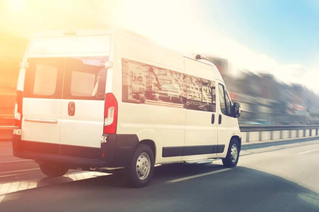 A white campervan driving down a highway, bathed in the warm glow of the setting sun. The vehicle's windows reflect the surrounding scenery, capturing a sense of adventure and freedom. The image conveys the excitement of road trips and exploring new destinations with the comfort and flexibility of a campervan.