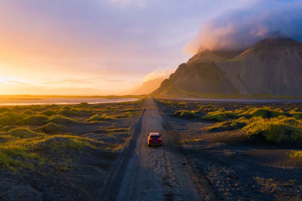 A bright mini car parked on a narrow road in Iceland. The landscape in the background shows vast, open fields with rugged mountains, showcasing the simplicity and practicality of exploring Iceland's natural beauty in a small, nimble car.