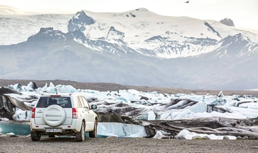 A white SUV parked near a glacial lagoon in Iceland. The vehicle faces a stunning landscape of icebergs floating in the water, with majestic snow-capped mountains in the background. The scene captures the rugged beauty of Iceland's natural wonders, emphasizing the adventure and exploration that awaits those who visit.