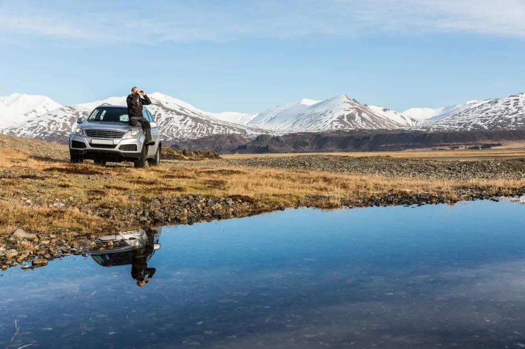 A person standing on the side of a parked SUV, holding binoculars and looking out at the scenic landscape. Snow-capped mountains rise in the background, reflected in a calm body of water in the foreground. The image captures the sense of adventure and exploration that comes with renting a car to explore Iceland's stunning natural beauty.