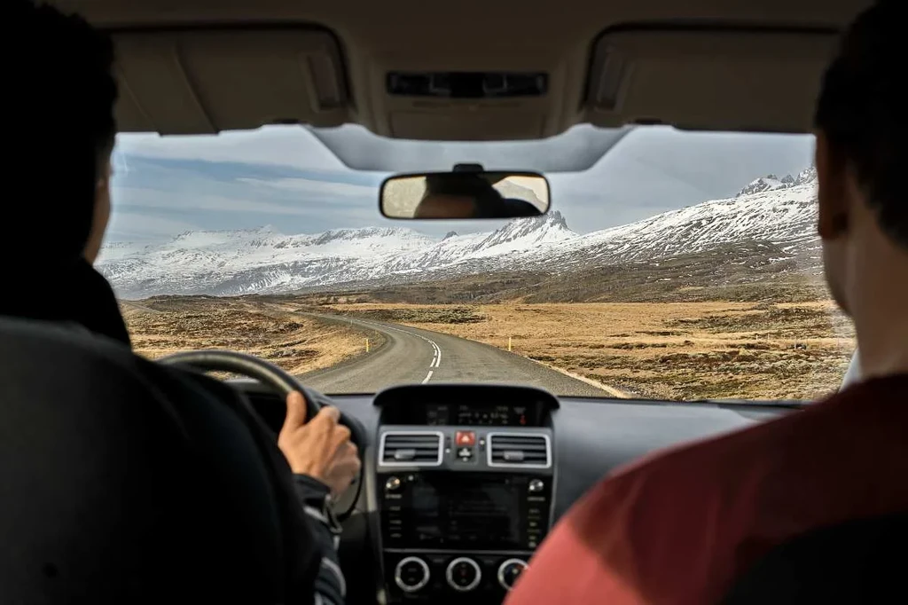 A view from the backseat of a car as it drives down a winding road in Iceland. The road stretches ahead, flanked by barren, brown fields leading to snow-covered mountains under a partly cloudy sky.