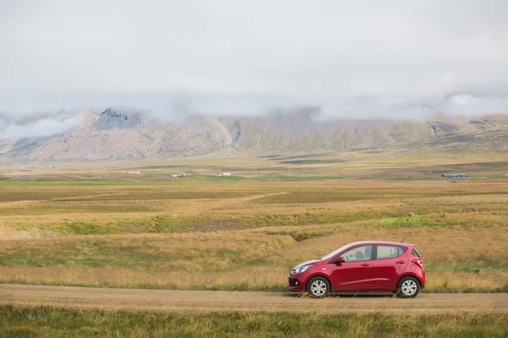 A small red car is parked on a dirt road in the foreground, with an expansive, grassy landscape stretching out behind it. The scene captures the vast, open plains of Iceland, with distant mountains partially shrouded in low-hanging clouds.