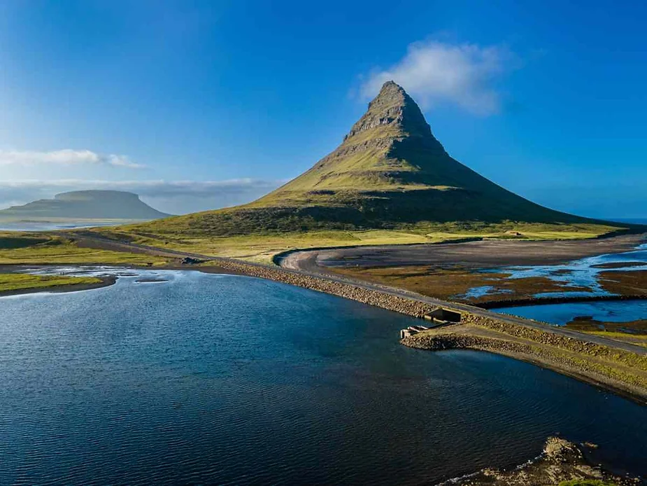 A picturesque landscape featuring a conical mountain surrounded by lush greenery and a winding road. A serene blue lake stretches in the foreground under a clear blue sky.