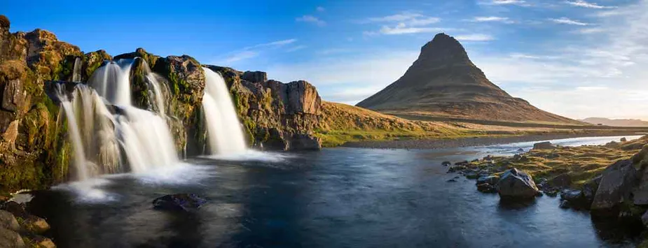 A stunning landscape showcasing a dual waterfall cascading into a tranquil river, surrounded by rugged rocks and vibrant greenery. A distinctively shaped conical mountain rises majestically in the background against a clear blue sky.