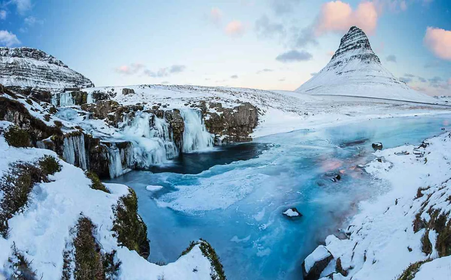 A stunning winter scene with a snow-covered conical mountain, frozen waterfall, and icy blue river. The landscape is blanketed in snow, with patches of green vegetation peeking through.