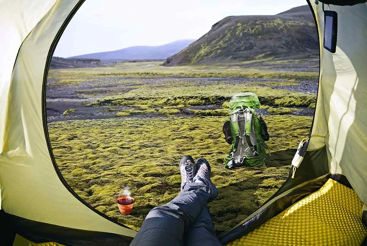 View from inside a tent in Iceland in July, showing a person's legs, a backpack set on lush green moss, and a cup of hot drink, with a mountainous landscape in the background.