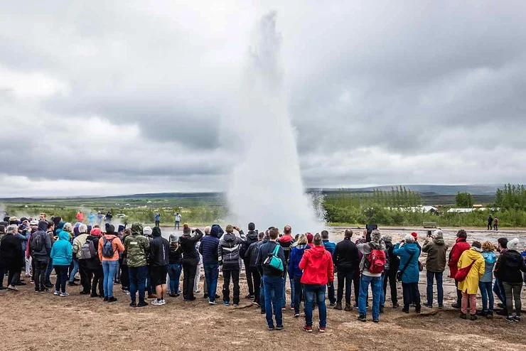 A crowd of people gathered to watch a geyser erupt in Iceland, with a tall plume of water shooting into the cloudy sky.